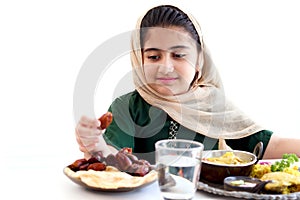 Adorable smiling Pakistani Muslim girl with beautiful eyes sitting at kitchen table, kid with hijab holding date fruit, enjoy