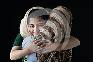 Adorable smiling Pakistani Muslim girl with beautiful eyes wearing hijab, hugging her mother on dark black background, warm love