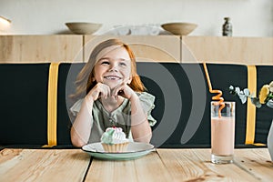 adorable smiling redhead girl with cupcake and milkshake sitting at table