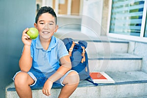 Adorable student boy smiling happy holding green apple sitting on the stairs at school