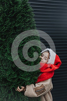 Adorable teen girl in Santa hat hugging Christmas tree outdoors. Girl against the background of a black fence with green fir trees