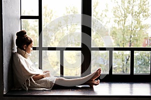 Adorable teenage girl sitting on a window sill, looking outside through the window.