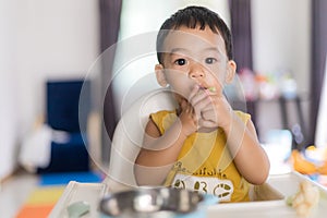 An Adorable toddler Asian baby boy 1-year-old sitting on a high feeding chair eating homemade food using hands and spoon at home