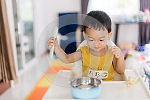 An Adorable toddler Asian baby boy 1-year-old sitting on a high feeding chair eating homemade food using hands and spoon at home