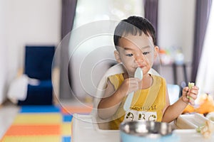 An Adorable toddler Asian baby boy 1-year-old sitting on a high feeding chair eating homemade food using hands and spoon at home