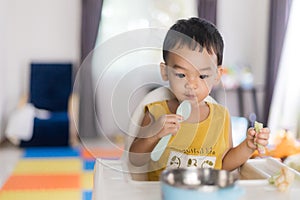 An Adorable toddler Asian baby boy 1-year-old sitting on a high feeding chair eating homemade food using hands and spoon at home