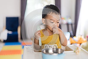 An Adorable toddler Asian baby boy 1-year-old sitting on a high feeding chair eating homemade food using hands and spoon at home