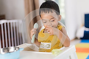 An Adorable toddler Asian baby boy 1-year-old sitting on a high feeding chair eating homemade food using hands and spoon at home