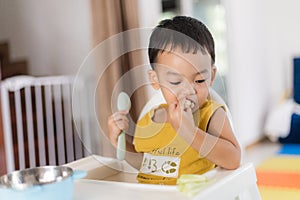 An Adorable toddler Asian baby boy 1-year-old sitting on a high feeding chair eating homemade food using hands and spoon at home