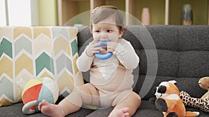 Adorable toddler bitting plastic hoop sitting on sofa at home