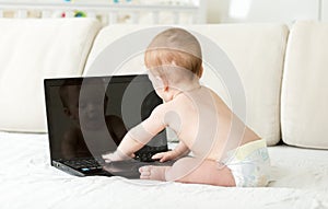 Adorable toddler boy in diapers sitting on sofa and typing message on laptop keyboard