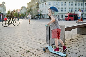 Adorable toddler boy riding his scooter in a city on sunny summer evening. Young child riding a roller