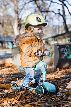 Adorable toddler boy wearing yellow protective helmet riding baby scooter outdoors on autumn day. Kid training balance