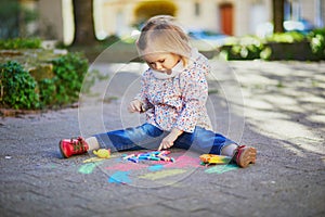 Adorable toddler girl drawing with colorful chalks on asphalt