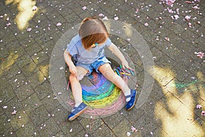 Adorable toddler girl drawing rainbow with colorful chalks on asphalt