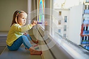 Adorable toddler girl drawing rainbow on window glass