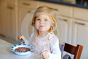 Adorable toddler girl eating healthy cereal with milk for breakfast. Cute happy baby child in colorful clothes sitting