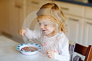 Adorable toddler girl eating healthy cereal with milk for breakfast. Cute happy baby child in colorful clothes sitting