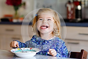 Adorable toddler girl eating healthy cereal with milk for breakfast. Cute happy baby child in colorful clothes sitting
