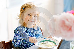 Adorable toddler girl eating healthy cereal with milk for breakfast. Cute happy baby child in colorful clothes sitting