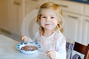 Adorable toddler girl eating healthy cereal with milk for breakfast. Cute happy baby child in colorful clothes sitting