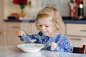 Adorable toddler girl eating healthy cereal with milk for breakfast. Cute happy baby child in colorful clothes sitting