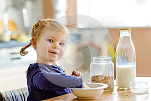 Adorable toddler girl eating healthy oatmeals with milk for breakfast. Cute happy baby child in colorful clothes sitting