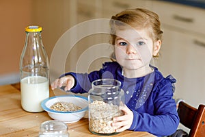 Adorable toddler girl eating healthy oatmeals with milk for breakfast. Cute happy baby child in colorful clothes sitting