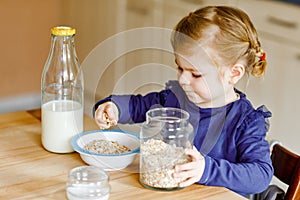 Adorable toddler girl eating healthy oatmeals with milk for breakfast. Cute happy baby child in colorful clothes sitting