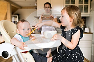 Adorable toddler girl at home feeding her baby brother.