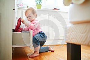 Adorable toddler girl at home, opening the drawer of dresser and taking out clothes