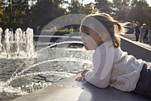 Adorable toddler girl looks at the pond with fountains in the park on a sunny day. weekend family walk. spending time with