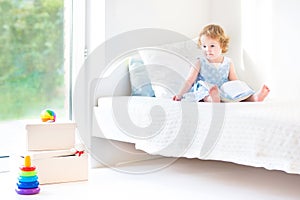 Adorable toddler girl reading book sitting on white bed