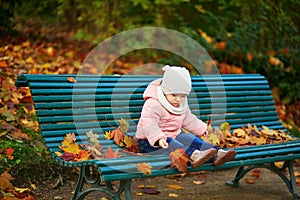 Adorable toddler girl sitting on the bench and playing with fallen leaves