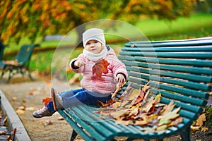 Adorable toddler girl sitting on the bench and playing with fallen leaves