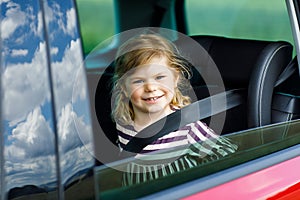 Adorable toddler girl sitting in car seat and looking out of the window on nature and traffic. Little kid traveling by