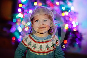 Adorable toddler girl sitting near Christmas tree. Little child playing and decorating xmas tree with family. First