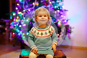 Adorable toddler girl sitting near Christmas tree. Little child playing and decorating xmas tree with family. First