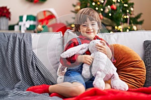 Adorable toddler hugging rabbit doll sitting on sofa by christmas tree at home