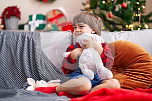 Adorable toddler hugging rabbit doll sitting on sofa by christmas tree at home