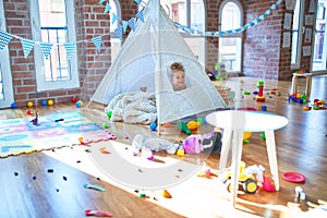 Adorable toddler smiling happy playing inside tipi around lots of toys at kindergarten