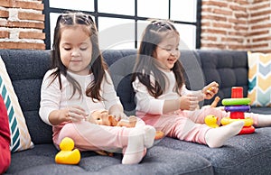 Adorable twin girls playing with hoops game holding baby doll sitting on sofa at home