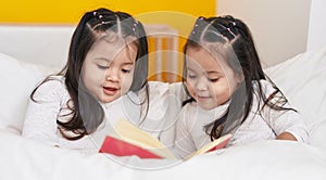 Adorable twin girls reading book sitting on bed at bedroom