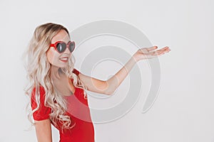 Adorable young blonde girl in red bodysuit and glasses in the shape of hearts posing on white background