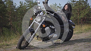 Adorable young caucasian woman in a black leather jacket and pants lying on a classic motorcycle. Hobby, traveling and