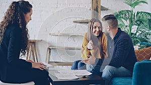 Adorable young couple house buyers are looking at papers and talking with realtor then smiling happily. Beautiful