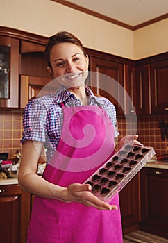 Adorable young european woman chocolatier in pink apron cute smiling while posing on camera with molds full of chocolate shells