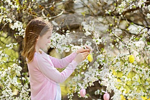 Adorable young girl having fun in blooming cherry garden on beautiful spring day. Kid hanging Easter eggs on blossoming cherry