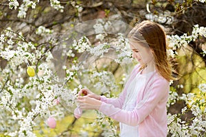 Adorable young girl having fun in blooming cherry garden on beautiful spring day. Kid hanging Easter eggs on blossoming cherry