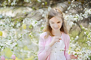 Adorable young girl having fun in blooming cherry garden on beautiful spring day. Kid hanging Easter eggs on blossoming cherry